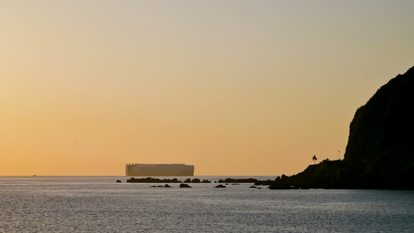 A large cargo ship sails on the calm ocean horizon during a golden sunrise, viewed from the rocky coast of Dawulun Beach in Keelung, Taiwan.