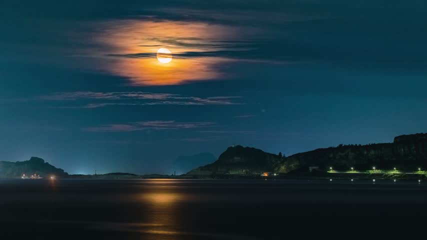 Full moon rising over the dark ocean casting a golden reflection on the water surface with distant coastal hills and night lights in Jinshan, Taiwan.