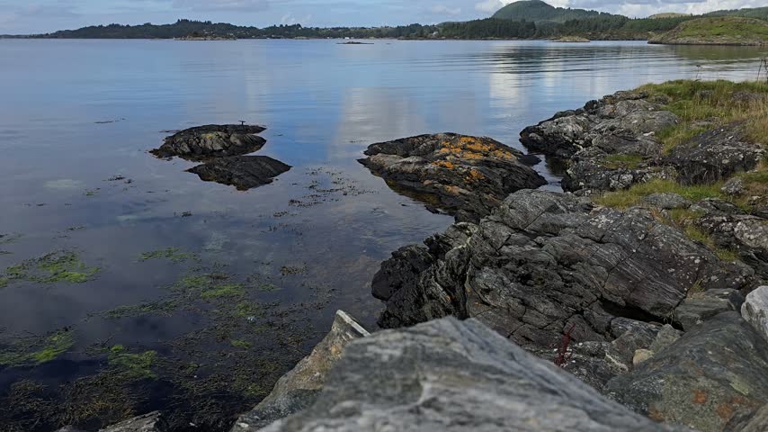 Large granite rocks dominate the foreground, while a dark forested shoreline with rolling hills stretches into the distance.Norway, Alver. September
