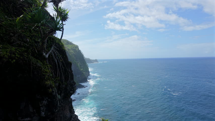 Dramatic green sea cliffs plunge into turquoise ocean water on Maui in Hawaii USA as white surf breaks over black volcanic rocks below a bright blue sky filled with drifting white clouds.