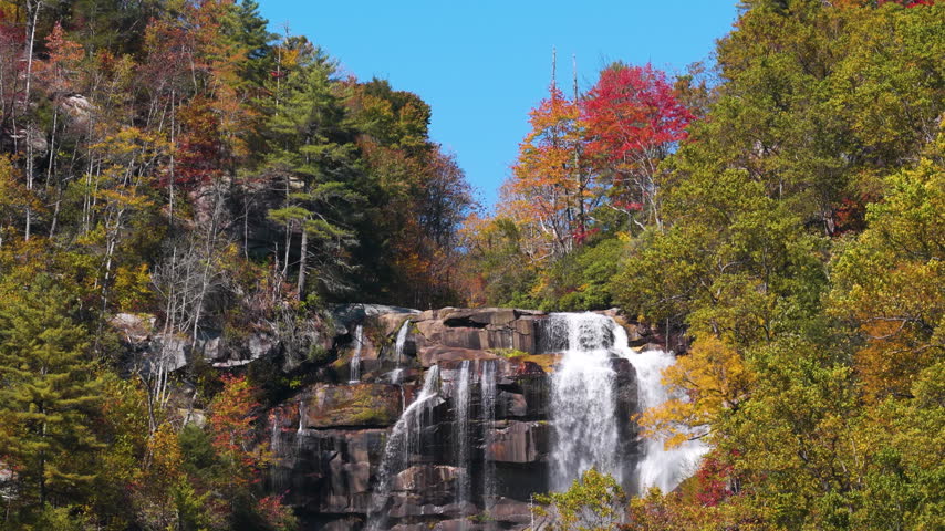 Majestic cascade pours through wooded mountain gorge surrounded by colorful autumn trees. Mist and rushing water create tranquil natural scenery.