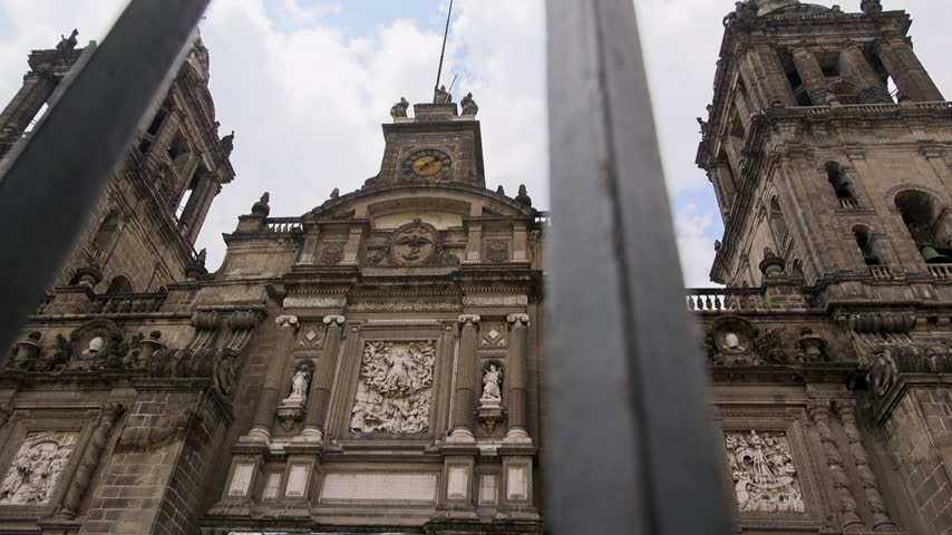 Low angle view of Mexico City Metropolitan Cathedrals main facade clock tower and statues seen through iron fence bars.