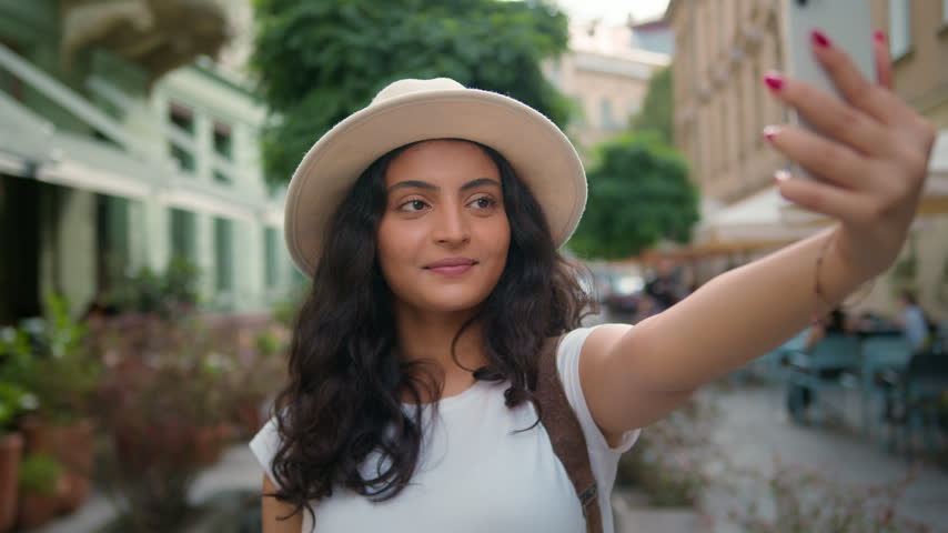 Happy young Indian woman taking selfie on mobile phone outside in city street Arabian ethnic female student tourist influencer use cellphone photo camera traveling vlog stream social media vacation