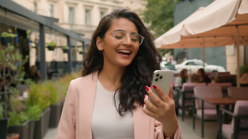 Happy young Indian woman making video call on mobile phone in city street cafe Arabian ethnic female girl businesswoman tourist talking greeting waving at camera business online connection app Wi-Fi