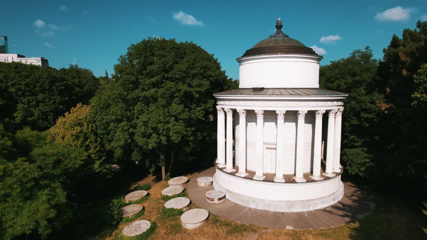A drone flies over a white neoclassical rotunda in a lush park, revealing the Warsaw city skyline in the background.