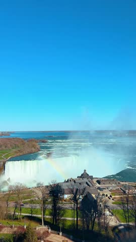 High angle view of Horseshoe Falls with rainbow at Niagara Falls. Water cascades into a misty basin under clear blue sky while a colorful spectrum forms over river.