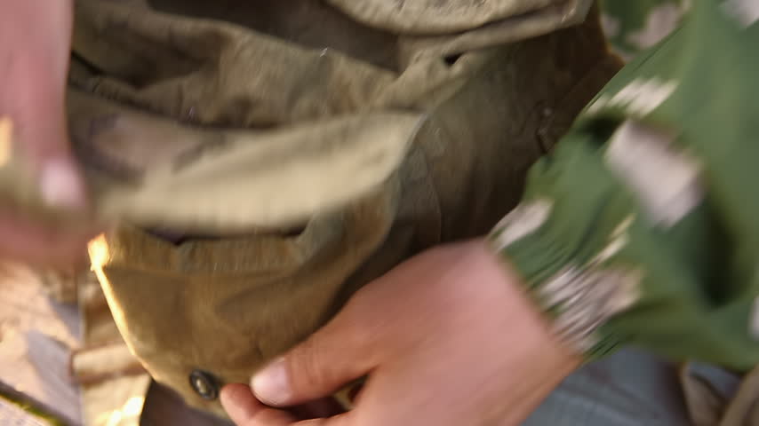 Close-up of a man in camouflage fastening the snap button on a canvas military backpack pocket.