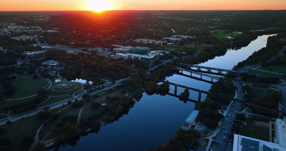 Footage over the Colorado River with bridges above it. Vast green cityscape of Austin, Texas, USA dazzled by the setting sun light.