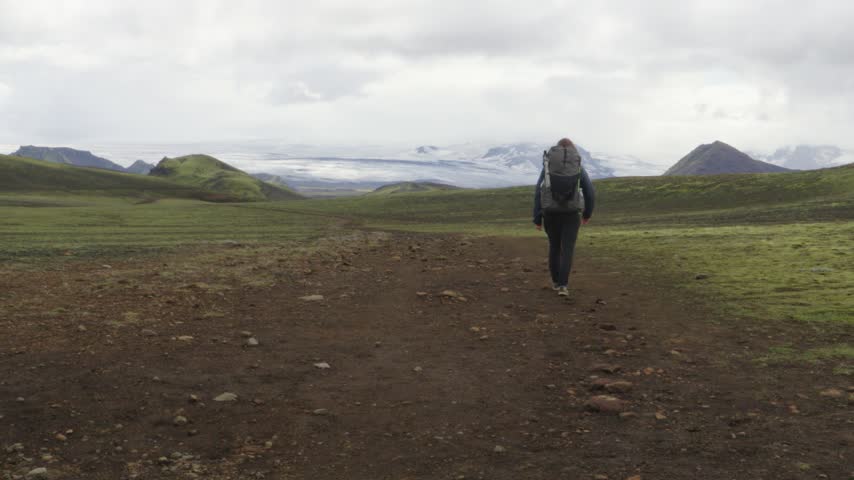 A lone hiker treks along the Laugavegur trail on Iceland, exploring the rugged volcanic landscape with distant glaciers under a cloudy sky. An iconic journey through nature
