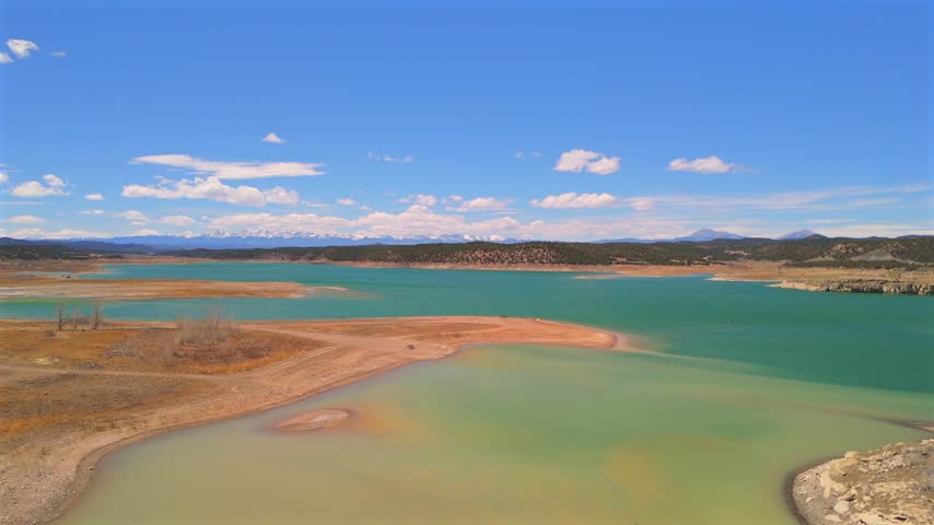 Drone glides above a curved sandy peninsula and shallow green inlet opening onto Trinidad Lake and distant Colorado mountain ranges