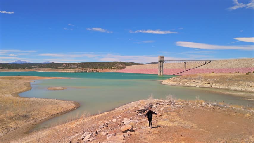 Rear aerial view follows a hiker in a wide-brimmed hat descending a rocky slope toward Trinidad Lake