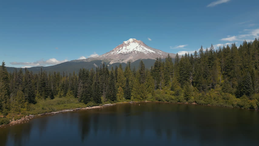 Mirror Lake and snow-capped Mount Hood rise beyond evergreen forest in Oregon, aerial medium ascend reveal behind treeline