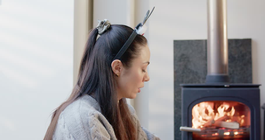 Adult woman in sweater sitting by stove watching fire, using scissors preparing gifts, copy space. Cozy, hearth, presents, wrapping, ribbon, warmth, home