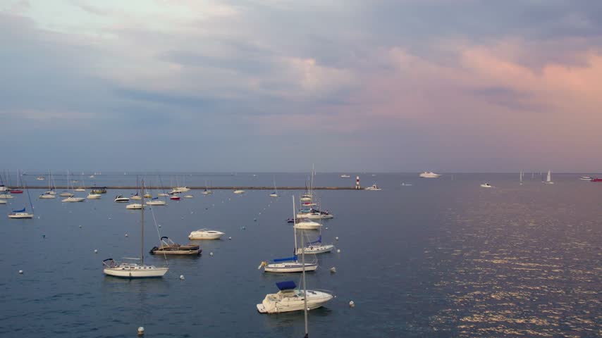 Wide drone view of sailboats and motorboats anchored in a Chicago harbor on Lake Michigan, with a breakwater lighthouse and soft pastel evening sky.