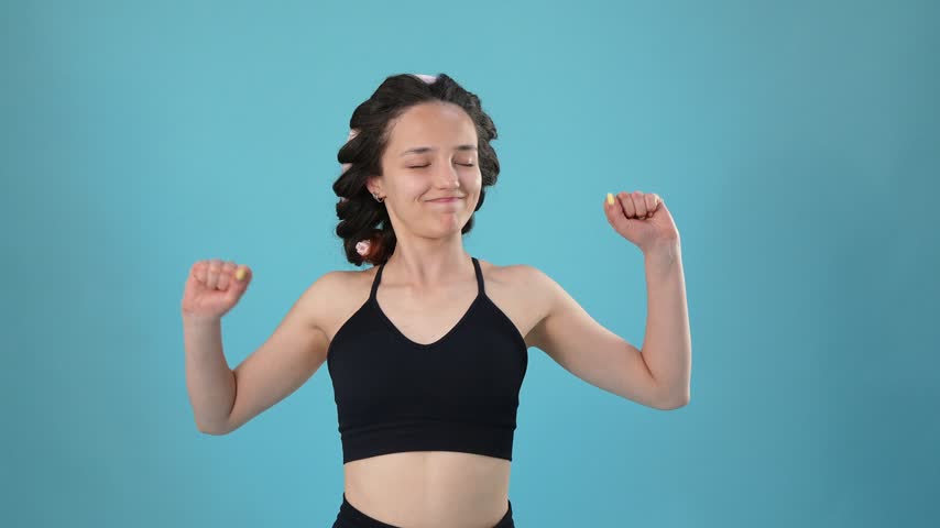 Young woman with heatless curlers in her hair stretches her arms and touches her face during a morning self-care routine against a bright blue background.