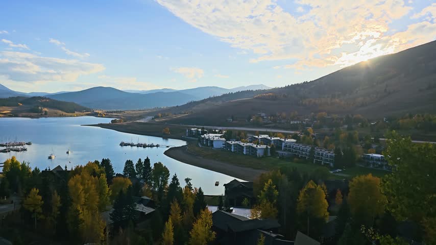Evening overlook of Dillon, Colorado, showing calm reservoir water, sailboats, lakeside condos, glowing fall foliage, and soft mountain silhouettes beneath dramatic sunlit clouds.