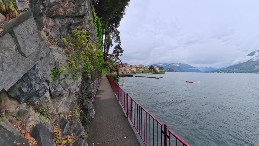 Scenic covered lakeside walkway with red metal arches beside a rocky garden wall, overlooking Lake Como and surrounding mountain scenery in Italy.