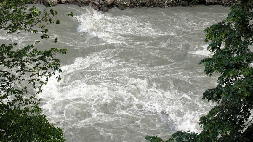 Top-down view of a powerful, wild mountain river with turbulent rapids and gray, muddy water flowing quickly through a rocky channel, framed by green foliage on the riverbanks