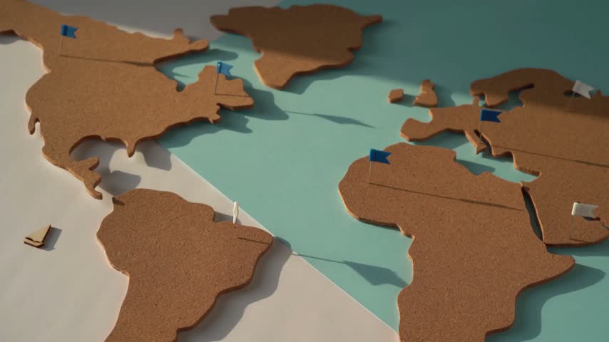 An aesthetic cork world map set against a two-toned background. A hand adorned with several silver rings places small wooden boat miniatures, one by one, onto the Atlantic Ocean area, creating a compelling visual representation of themes such as navigation, exploration, global adventure, and travel planning.