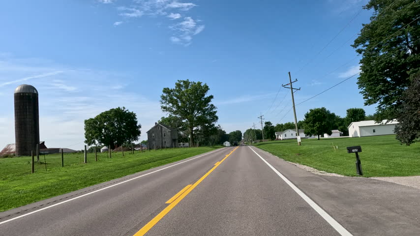 Car POV Driving Past Farmhouse and Cornfield in Rural Illinois. Car POV shot traveling along an asphalt highway past farmhouses, green cornfields, and quiet residential homes near Lebanon, Illinois, USA.
