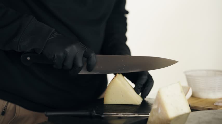 Skilled cheesemonger wearing black gloves slicing a wheel of aged goat cheese. Professional working in a dairy farm preparing a gourmet food sample