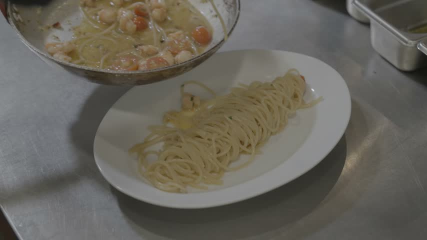 Close up chef plating shrimp spaghetti pasta onto white plate in kitchen.