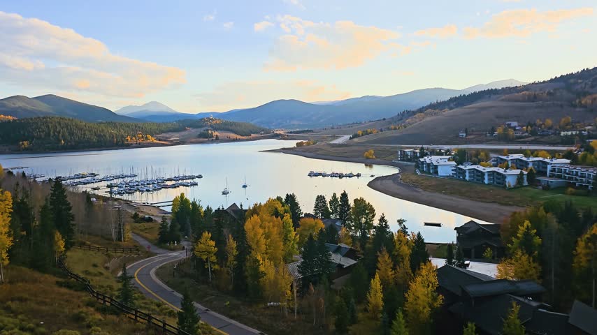 Scenic Dillon, Colorado overlook with blue reservoir water, colorful fall foliage, resort buildings, boat marina, curving road, and distant mountains in warm afternoon light.