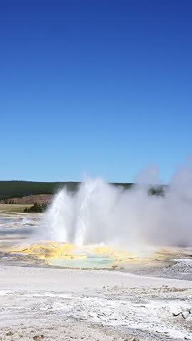 Clepsydra Geyser erupting with yellow minerals at base and small hot spring, sunny blue sky day, summer vacation at Yellowstone National Park viewing natural geothermal features
