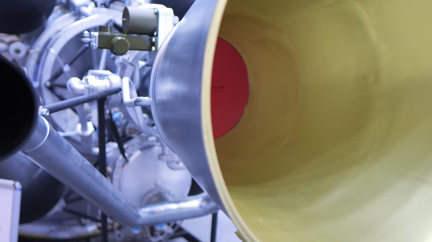 Close-up of rocket engine nozzle and mechanical components. Metal pipes and gears visible. Yellow interior of engine chamber. Industrial machinery background