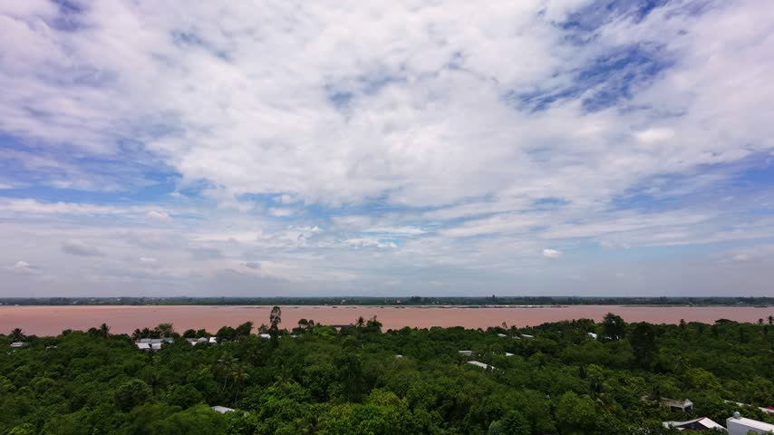 A high-angle aerial drone shot capturing a distant view of the expansive Mekong River flowing along the horizon. In the foreground, a dense canopy of green orchards and scattered local rooftops stretches out beneath a vast, cloudy blue sky, emphasizing the impressive breadth of the silt-laden river and the surrounding rural countryside.