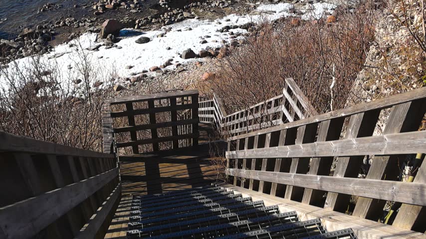 High angle view descending a long wooden staircase down a cliff to the rocky shore of the Baltic Sea, where patches of snow remain on the beach during a sunny early spring day in Paldiski