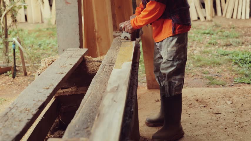 Carpenter operates an electric wood planer to smooth a wooden plank at an outdoor rural sawmill