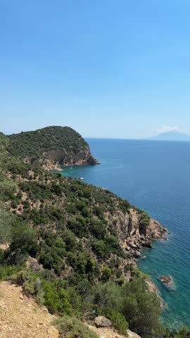 Scenic view of rocky cliffs and green coastal hills on Thasos Greece with a sailboat in a turquoise cove and a distant mountain on the horizon beneath a clear blue sky
