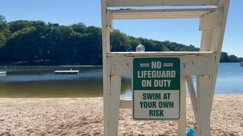 A green no lifeguard on duty sign is attached to an empty white chair with an empty plastic cup blowing on top near a lake beach.