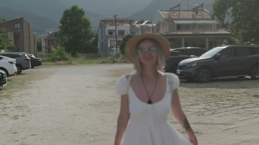 A blonde woman in mirrored sunglasses and a straw hat turns from side to side and smiles while holding her white dress near her shoulder against a blurred street background