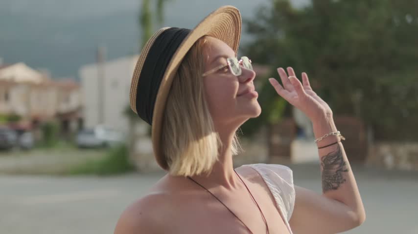 A blonde woman in mirrored sunglasses and a straw hat turns from side to side and smiles while holding her white dress near her shoulder against a blurred street background