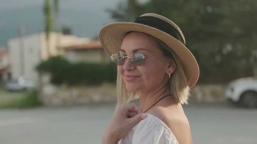 A blonde woman in mirrored sunglasses and a straw hat turns from side to side and smiles while holding her white dress near her shoulder against a blurred street background