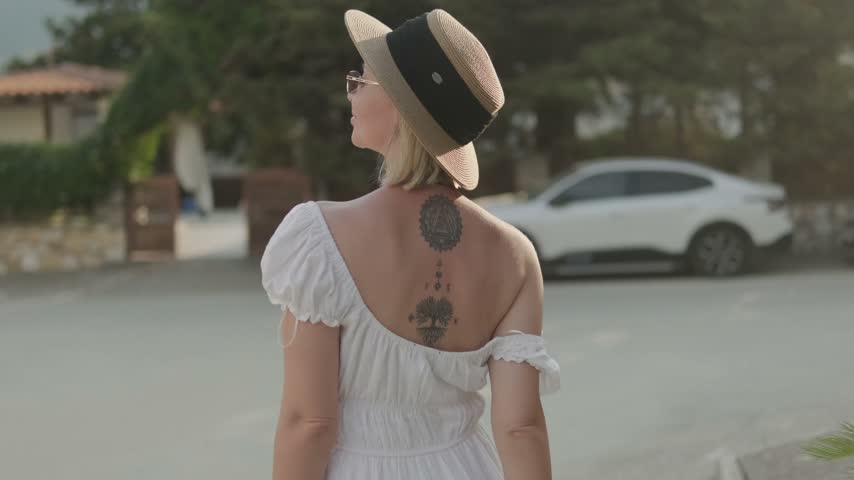 A blonde woman in mirrored sunglasses and a straw hat turns from side to side and smiles while holding her white dress near her shoulder against a blurred street background
