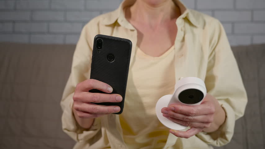 Woman connecting smart home security camera to smartphone. Woman holding a modern wireless security camera and a smartphone, demonstrating the process of connecting and configuring the device