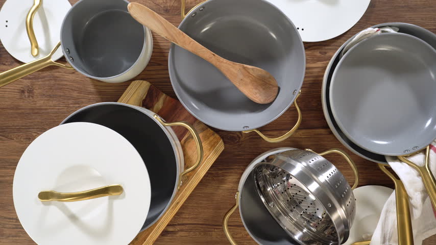 A top-down view shows a collection of non-stick pots, pans, and lids with golden handles arranged neatly on a wooden table. The scene is bright and clean, emphasizing modern kitchenware.