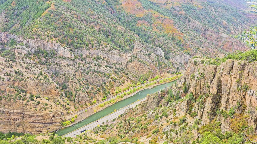 High overlook reveals a winding river and highway through a rugged mountain canyon, with steep rock cliffs, evergreen forest, autumn foliage, and scenic transportation routes below.