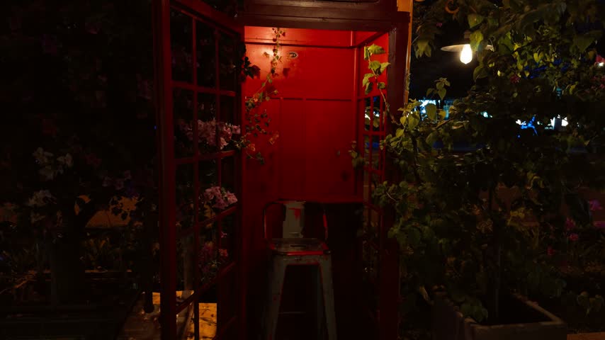 Red telephone booth glowing brightly at night, transformed into a decorative element with plants and a metal stool, creating a romantic and moody ambiance on a da nang street