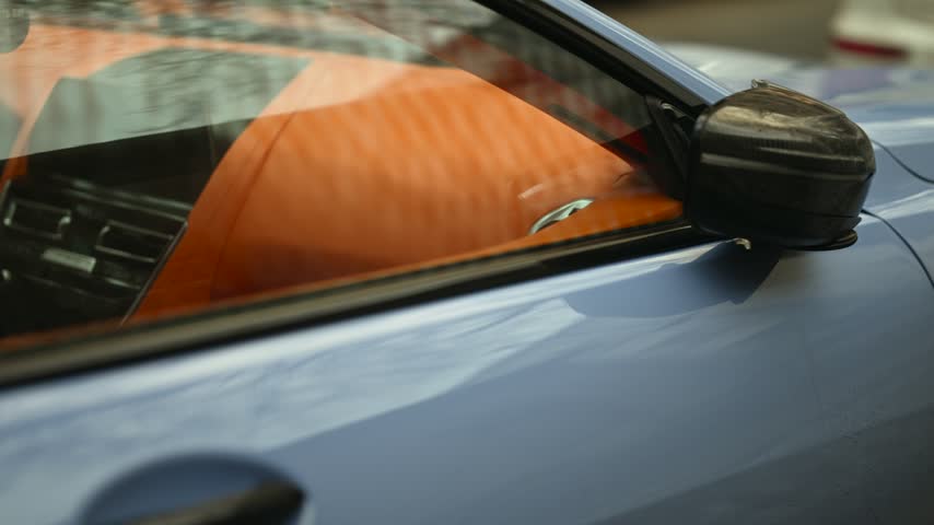Blue sports car exterior close up focusing on carbon fiber side mirror housing and vibrant orange leather dashboard seen through clean window glass during daylight outdoor scene.