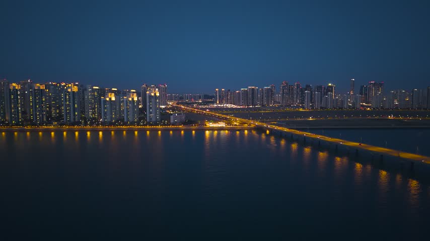 City lights illuminate Songdo skyline and curved bridge reflecting on calm water as evening traffic crosses causeway toward apartment towers