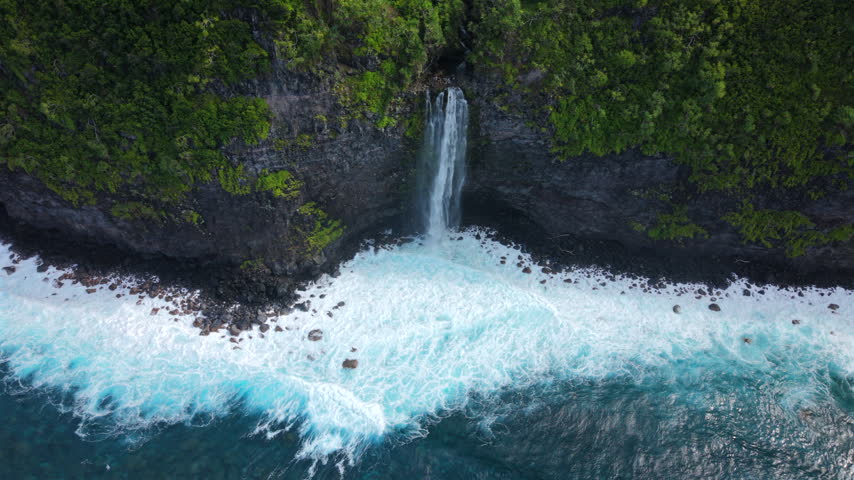 Aerial top down view of a secret waterfall on Maui Hawaii USA plunging from a lush green sea cliff into foaming turquoise ocean surf breaking over dark volcanic rocks along wild tropical shore.