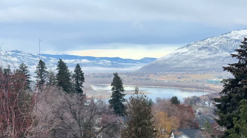 Handheld shot of a serene winter landscape at Mount Paul, Kamloops, featuring snow-covered mountains, a river, and a town nestled between the hills, framed with trees in the foreground, balanced co...