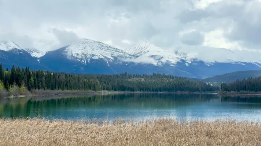 Static shot of a serene lake reflecting snow-capped mountains and evergreen forest under a cloudy sky at Jasper National Park, Canada.