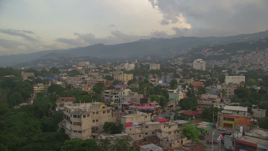 View from above of densely packed concrete buildings and lush trees in Port-au-Prince, Haiti. Urban sprawl nestled against dramatic mountain ranges under moody skies.