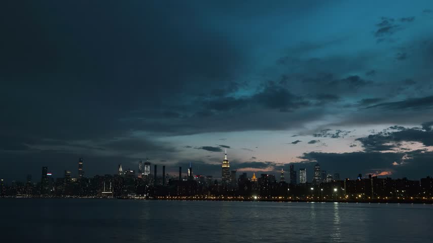 A wide panoramic view of the Manhattan skyline at twilight, with city lights reflecting on the water under a dramatic stormy sky.