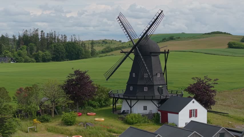 Drone footage circling a charming countryside farm and its traditional windmill, revealing the vibrant green meadows and idyllic rural landscape of Eastern Jutland, Denmark.